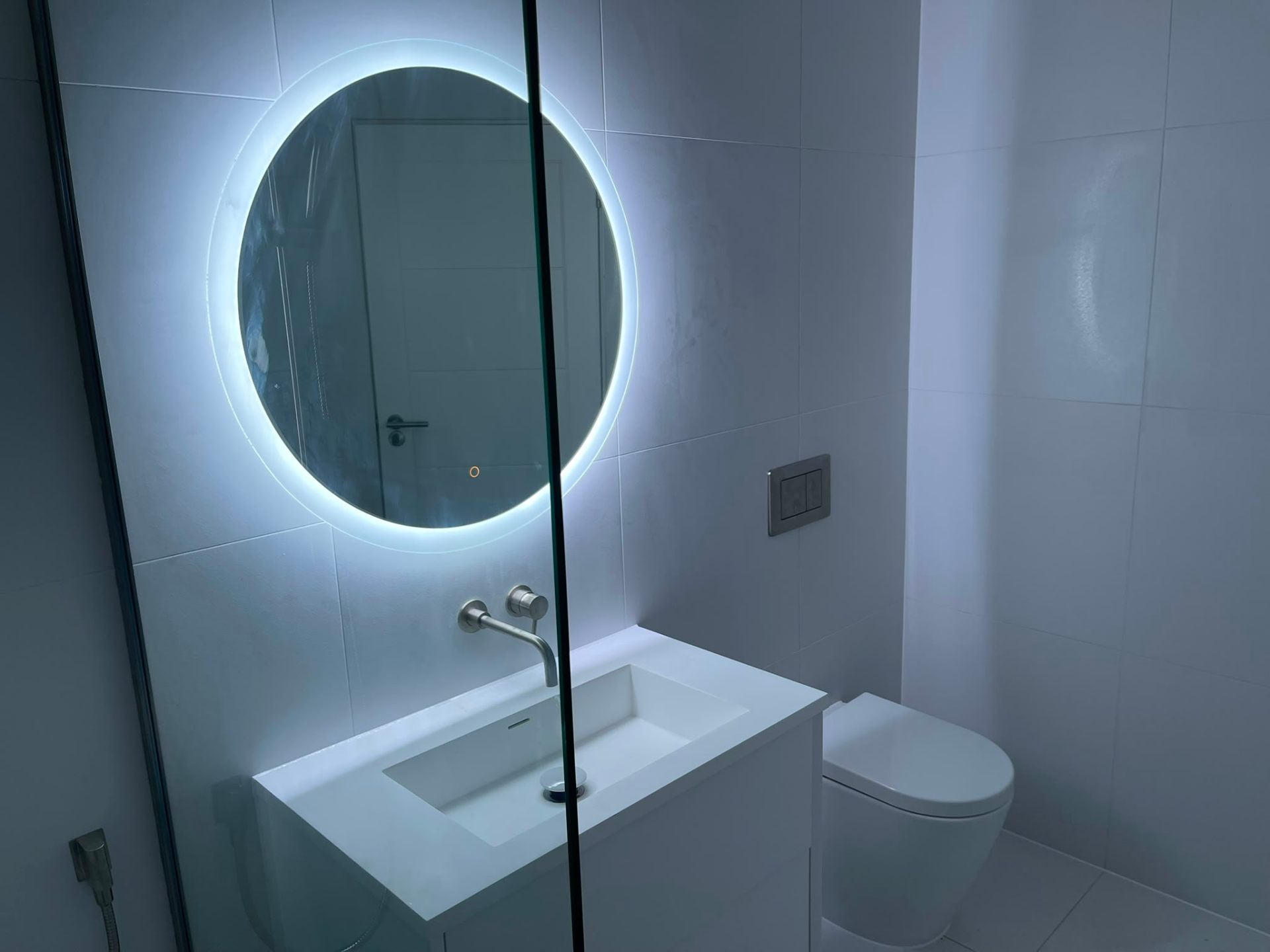 White cloakroom with backlit round mirror, wall-hung basin and WC by Heat Solution London